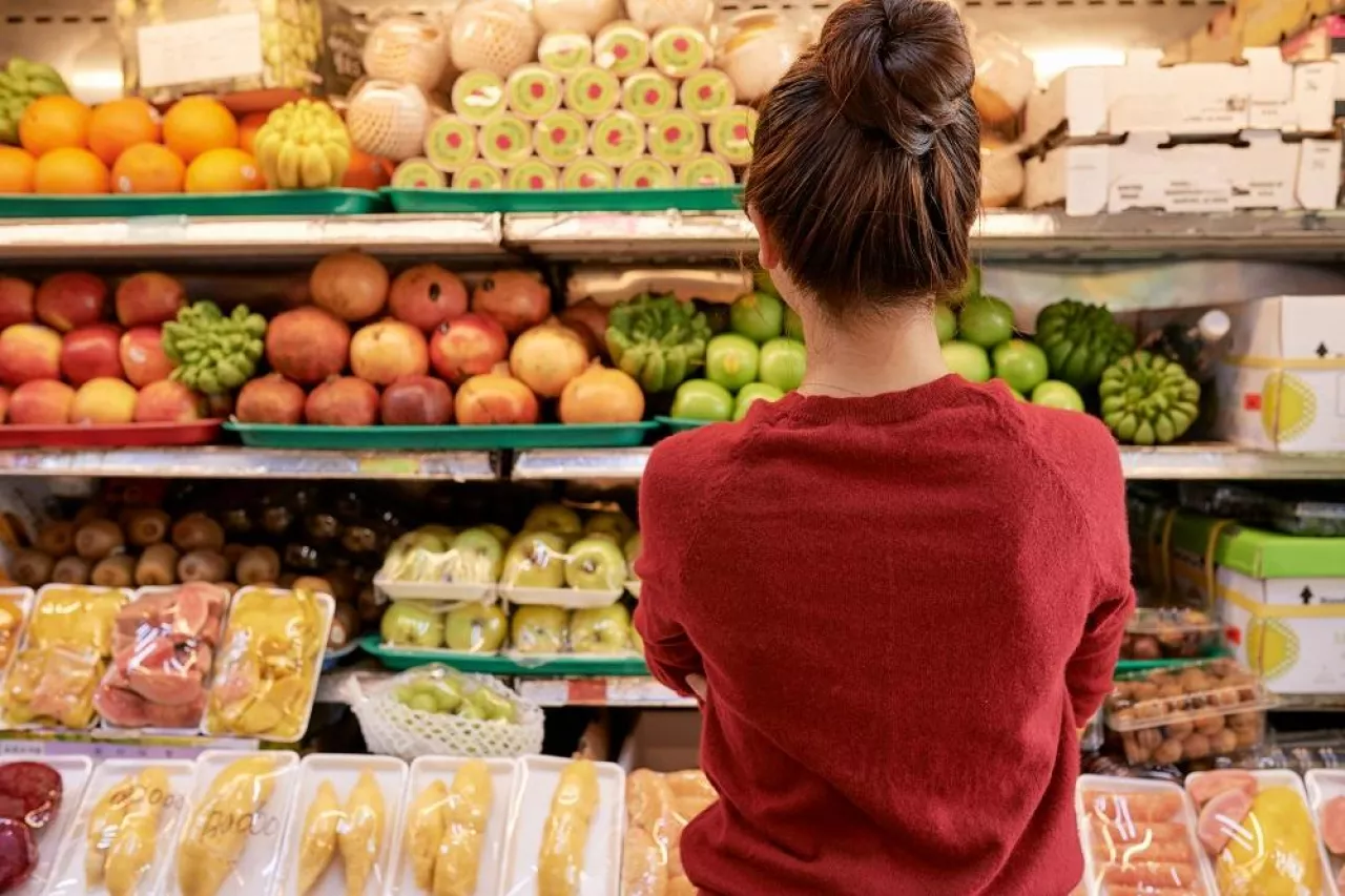 Pensive young woman looking at shelves with fresh fruits and vegetables at supermarket