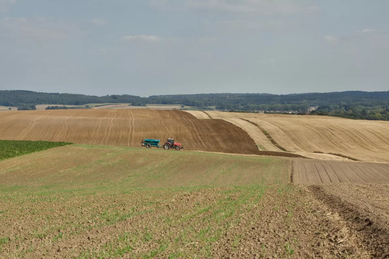 Ciągnik jadący z zaczepionym rozrzutnikiem nawozów wśród pól rolniczych.