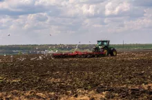 Zielony ciągnik marki John Deere, agregatowany z czerwoną broną talerzową, wykonuje prace uprawowe na polu. Scena ukazuje klasyczny etap przygotowania stanowiska pod zasiewy w warunkach optymalnej wilgotności gleby.